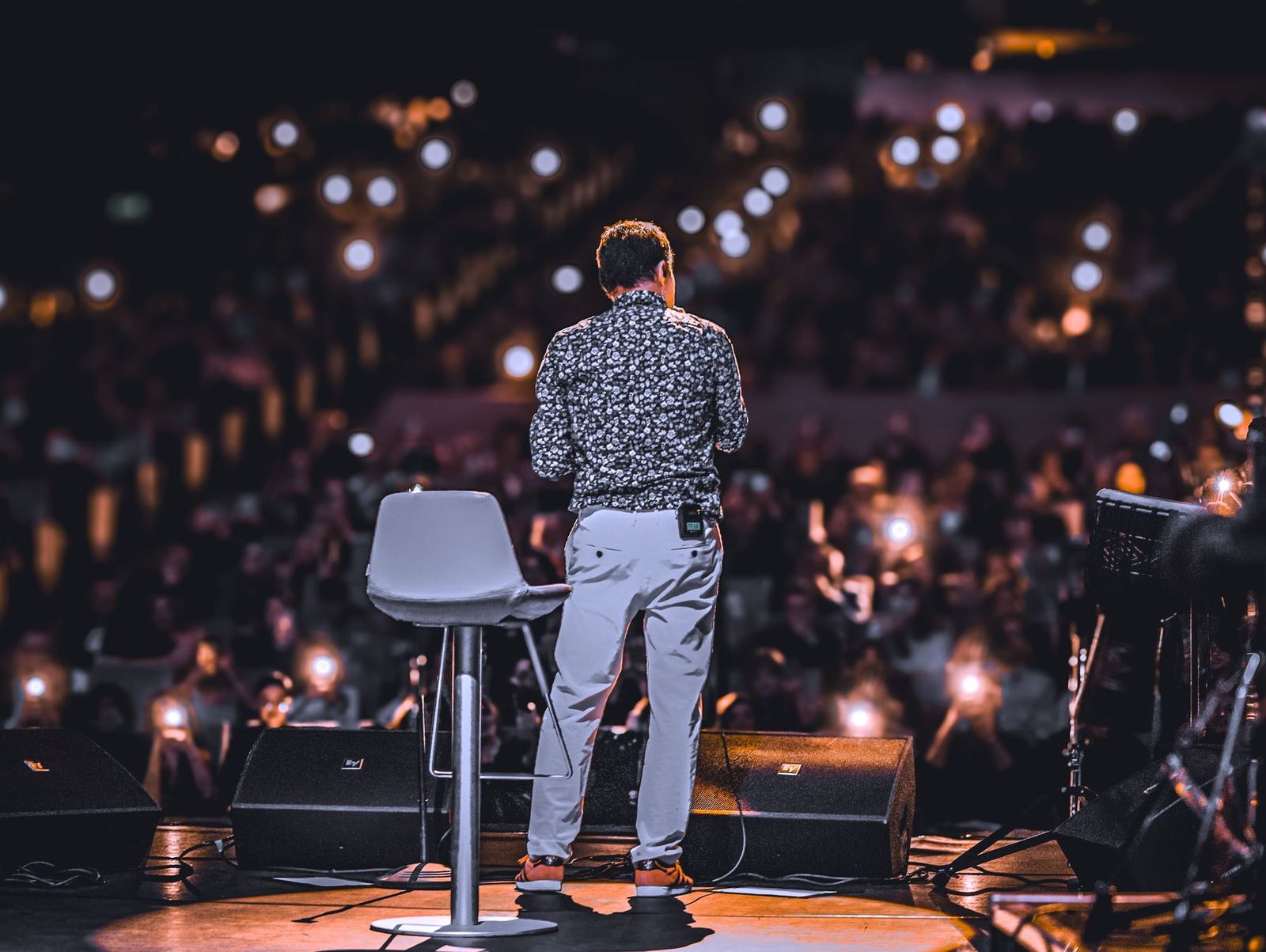 Palestrante de costas no palco, diante de uma plateia enorme com as luzes dos celulares acesas