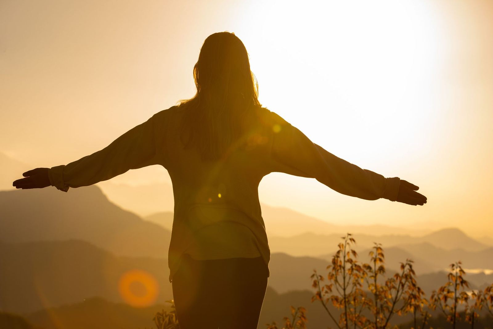 Mulher de braços abertos no alto de um morro, contra o sol nascendo sobre as montanhas