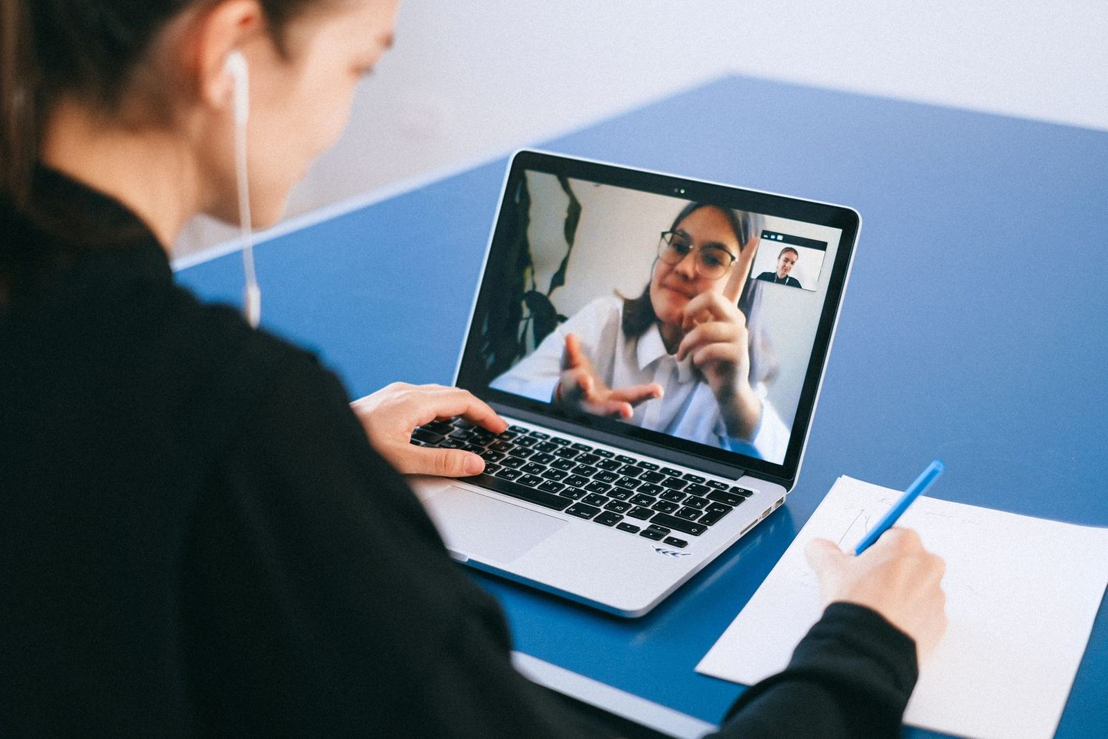Aluna diante do notebook numa aula ao vivo por vídeo, anotando no papel enquanto a professora fala na tela