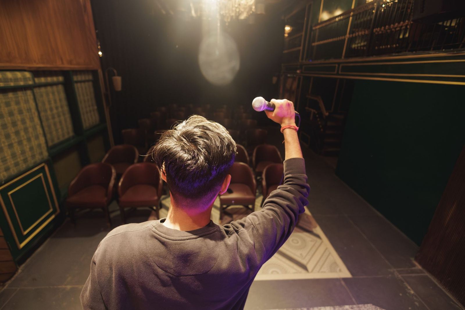 Rapaz de costas no palco de um teatro, erguendo o microfone para a plateia