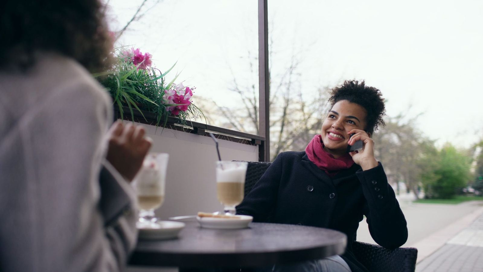 Mulher sorrindo ao telefone, sentada à mesa de um café na calçada