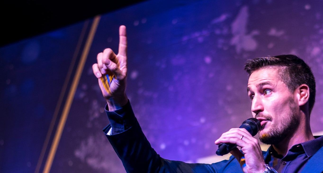 Victor Reis no palco, microfone na mão e o dedo erguido enquanto fala